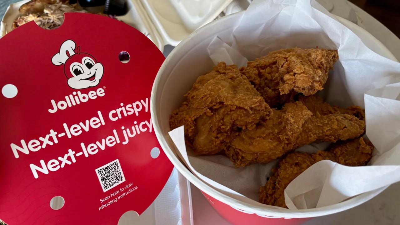 Fried chicken pieces in a paper-lined bucket beside its red Jollibee lid, which reads Next-level crispy, Next-level juicy, with a QR code for reheating instructions