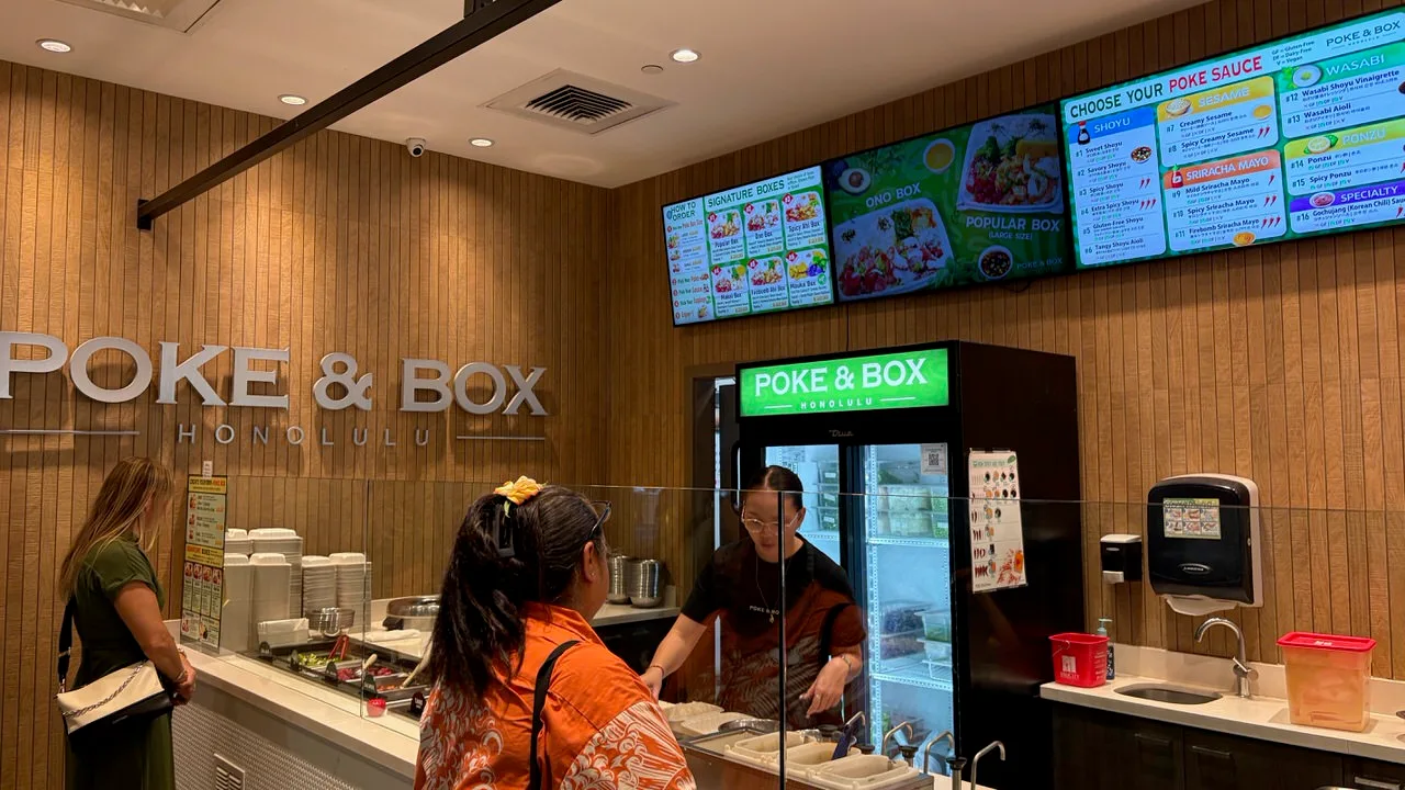 The Poke & Box counter with HONOLULU lettering on a wood-slat wall, a green-lit drinks fridge, and screens showing signature boxes and a numbered list of poke sauces