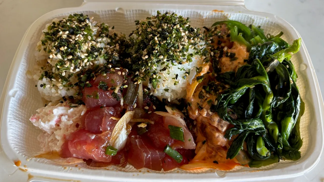 A poke box with cubes of raw ahi, green onion and onion, ahi in an orange spicy mayo, two scoops of rice under furikake and sesame, and dark cooked greens