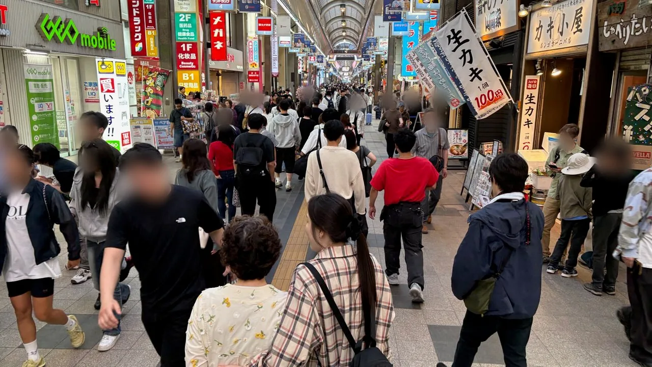 A covered shopping arcade full of people walking, with shop signs and a banner advertising raw oysters at 100 yen each