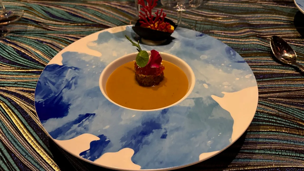 A blue and white plate holding a round mound of crab topped with orange roe and a leaf, sitting in orange lobster bisque, with a small black boat-shaped dish and red coral garnish behind