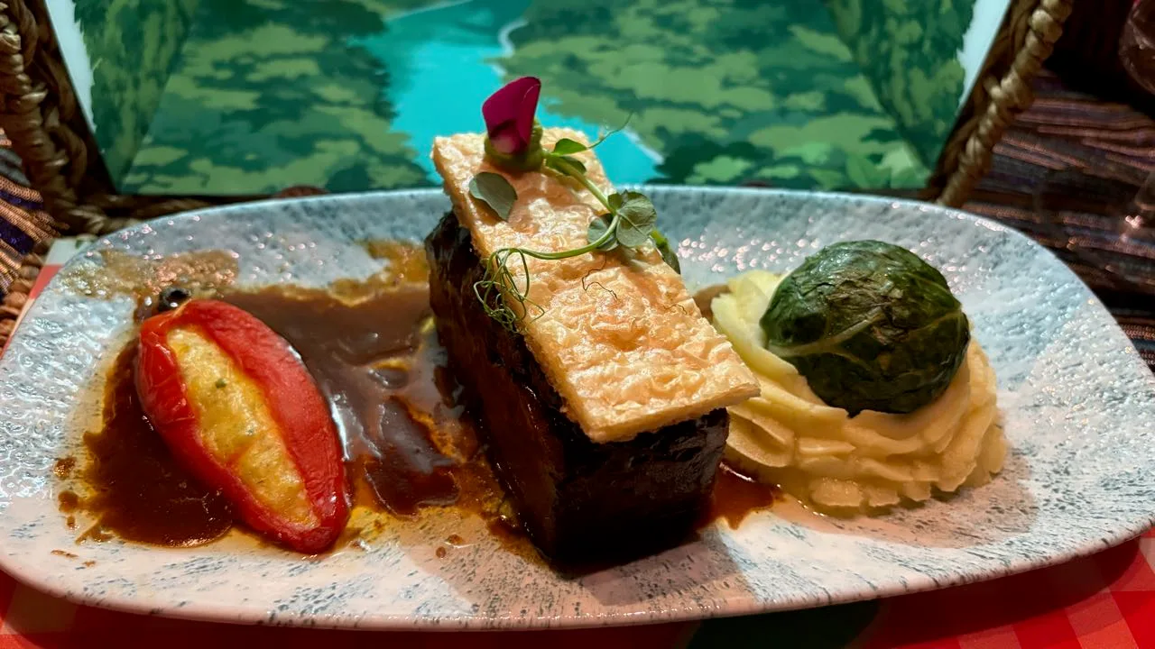A block of dark braised short rib with a flat rectangle of puff pastry laid on top, a stuffed red pepper in brown sauce and mashed potato under a cabbage parcel