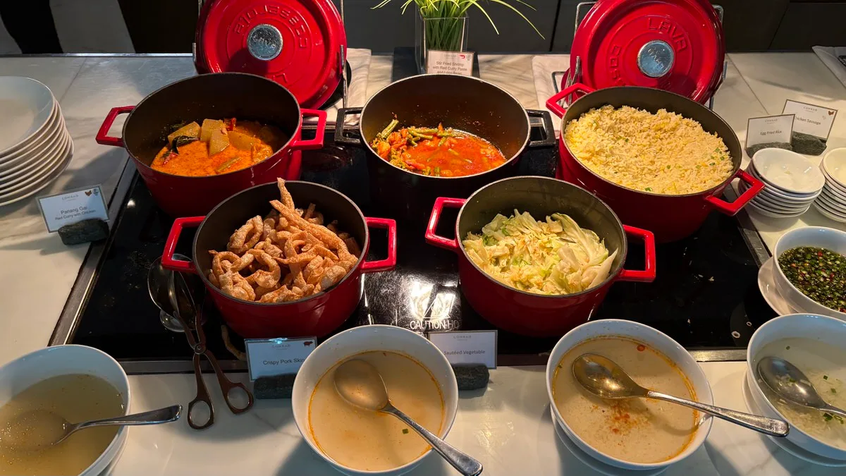 Red cast-iron pots on a buffet counter holding a red chicken curry, stir-fried shrimp with long beans, egg fried rice, crispy pork skin and sautéed cabbage, with bowls of soup and a dish of chili sauce in front