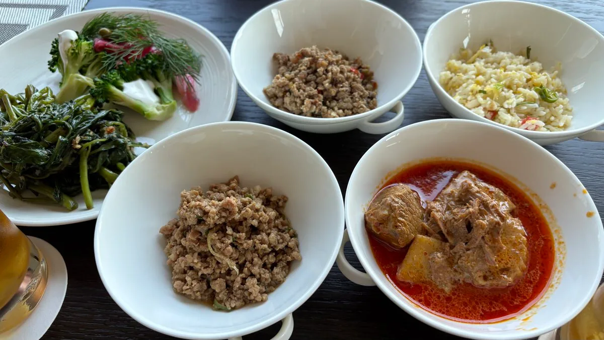 A plate of stir-fried morning glory and broccoli with a sprig of feathery green herb and pieces of red salmon, beside four bowls: two of minced meat, one of fried rice and one of beef curry in a red-orange sauce