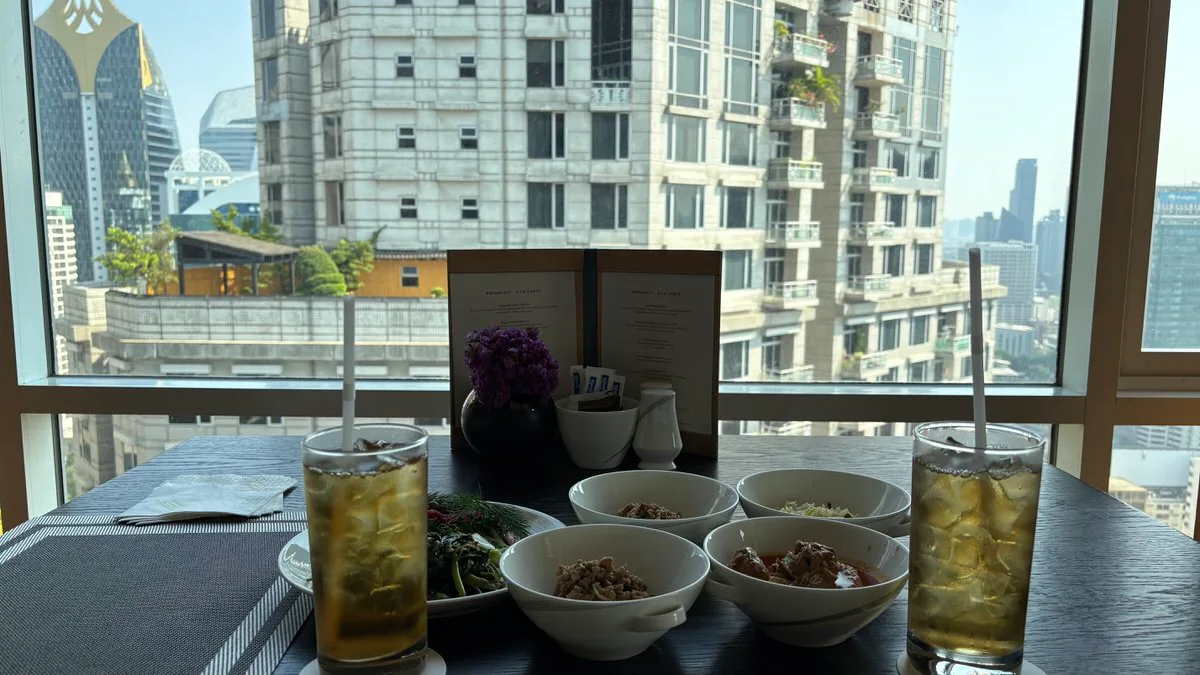Two tall glasses of iced tea with straws on a dark table by a window, with bowls of food, a plate of greens, a standing menu and a vase of purple flowers, and city buildings outside