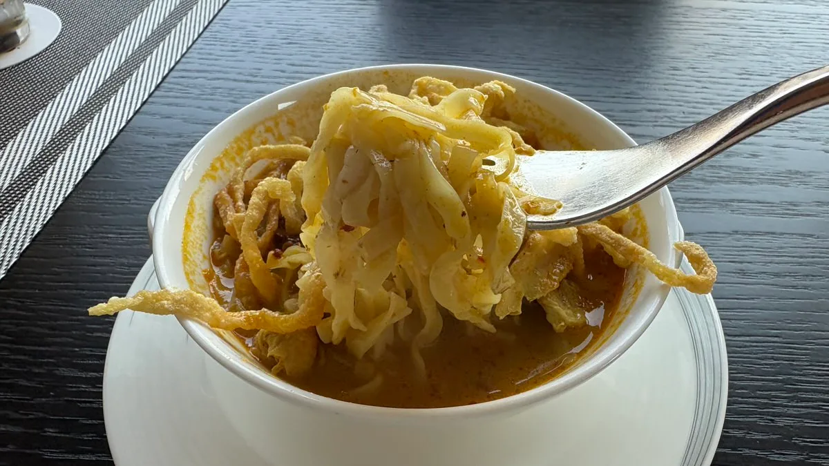 A fork lifting soft yellow noodles out of a white bowl of curry broth, with crispy fried noodles around the edge of the bowl