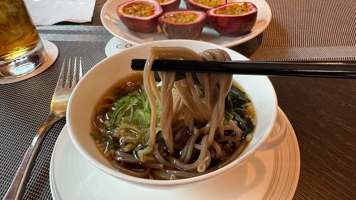 Black chopsticks lifting grey-brown soba noodles from a white bowl of dark broth with green onion, wakame and tempura bits, and a plate of halved passion fruit behind