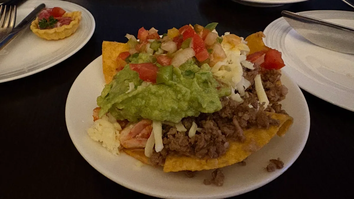 Tortilla chips on a white plate under minced beef, grated cheese, a mound of guacamole and diced tomato salsa, with a small tart on a plate behind