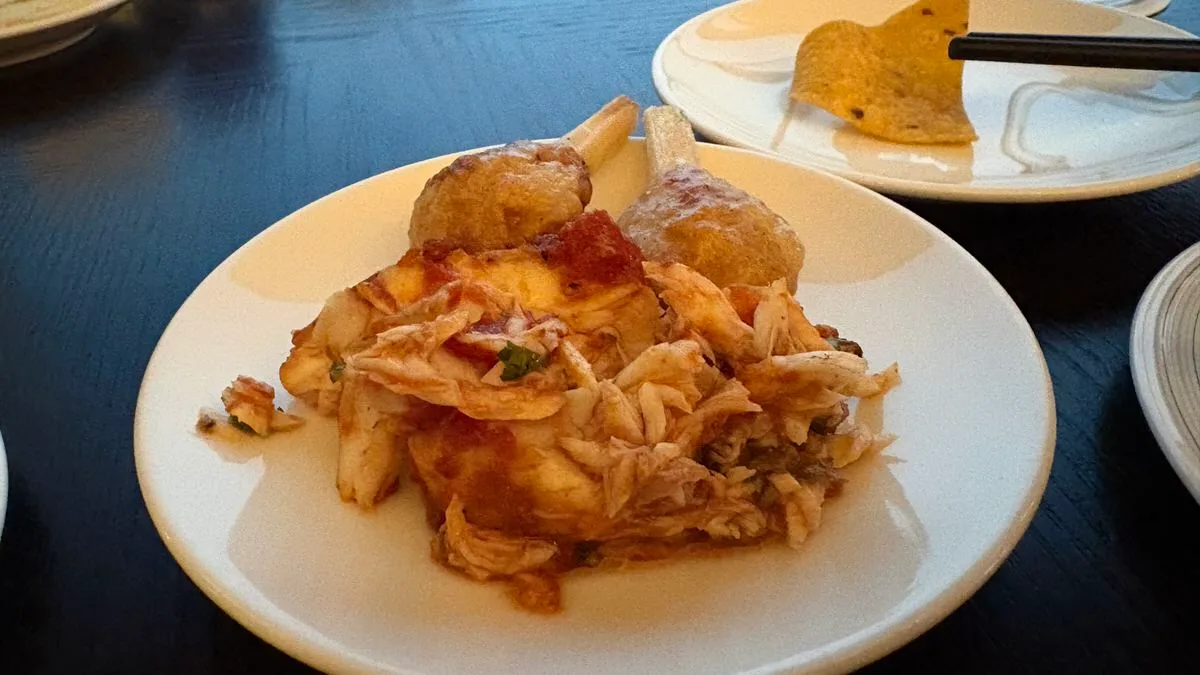 Flaked fish in a red sauce on a white plate, two fried shrimp balls on sugarcane sticks behind it, and a tortilla chip on another plate at the back