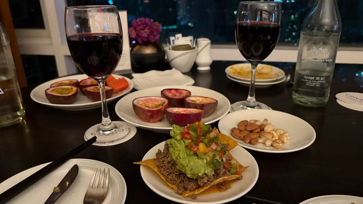 Two glasses of red wine on a dark table, two plates of halved passion fruit, a small plate of almonds and cashews, and a tortilla chip topped with beef, guacamole and diced salsa