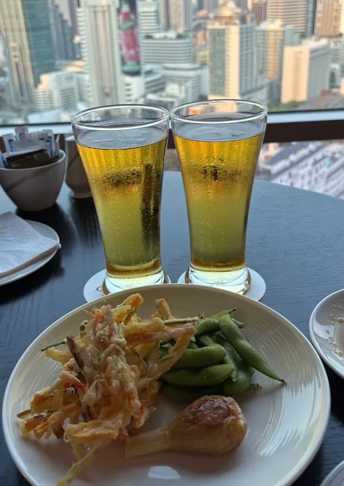 Two glasses of beer on a dark table by the window with city towers behind, and a plate of vegetable tempura, edamame and a fried shrimp ball on a sugarcane stick