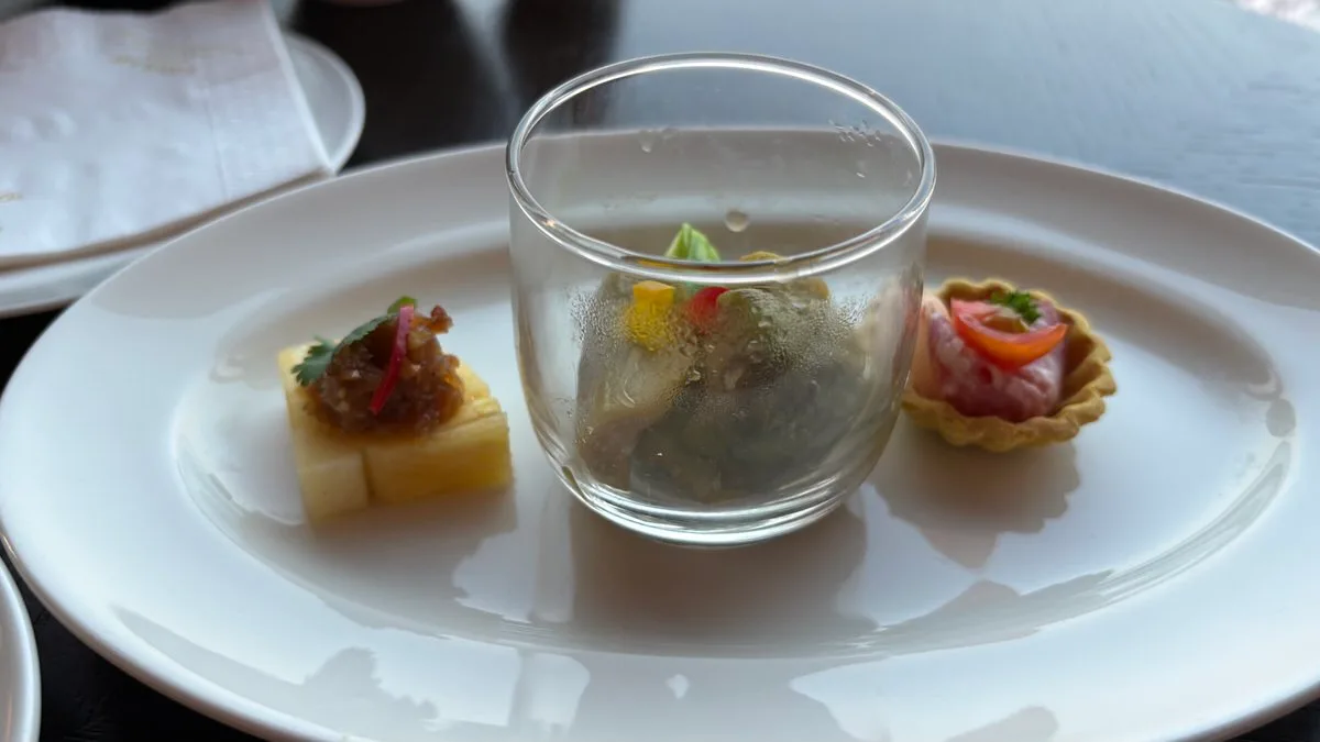 Three canapés on a white plate: a yellow cube topped with a brown relish, red onion and a coriander leaf, a small glass cup of green vegetables, and a pastry tart with a pink topping and a slice of tomato