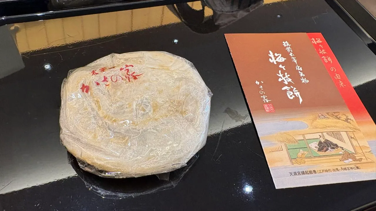 A single plastic-wrapped umegae-mochi with red Kasa no Ya lettering on a black tray, beside an illustrated Kasa no Ya leaflet