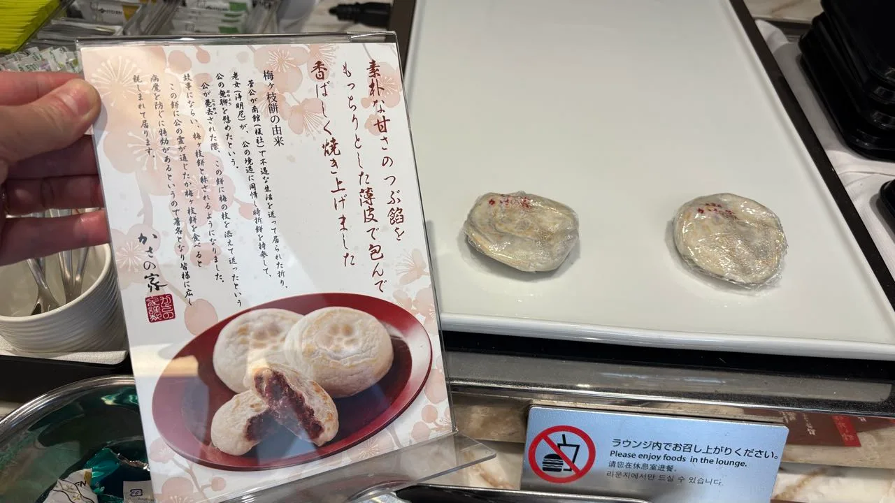 A hand holding up a Kasa no Ya display card with a photo of umegae-mochi, beside two plastic-wrapped umegae-mochi on a white tray and a sign asking guests to eat food in the lounge