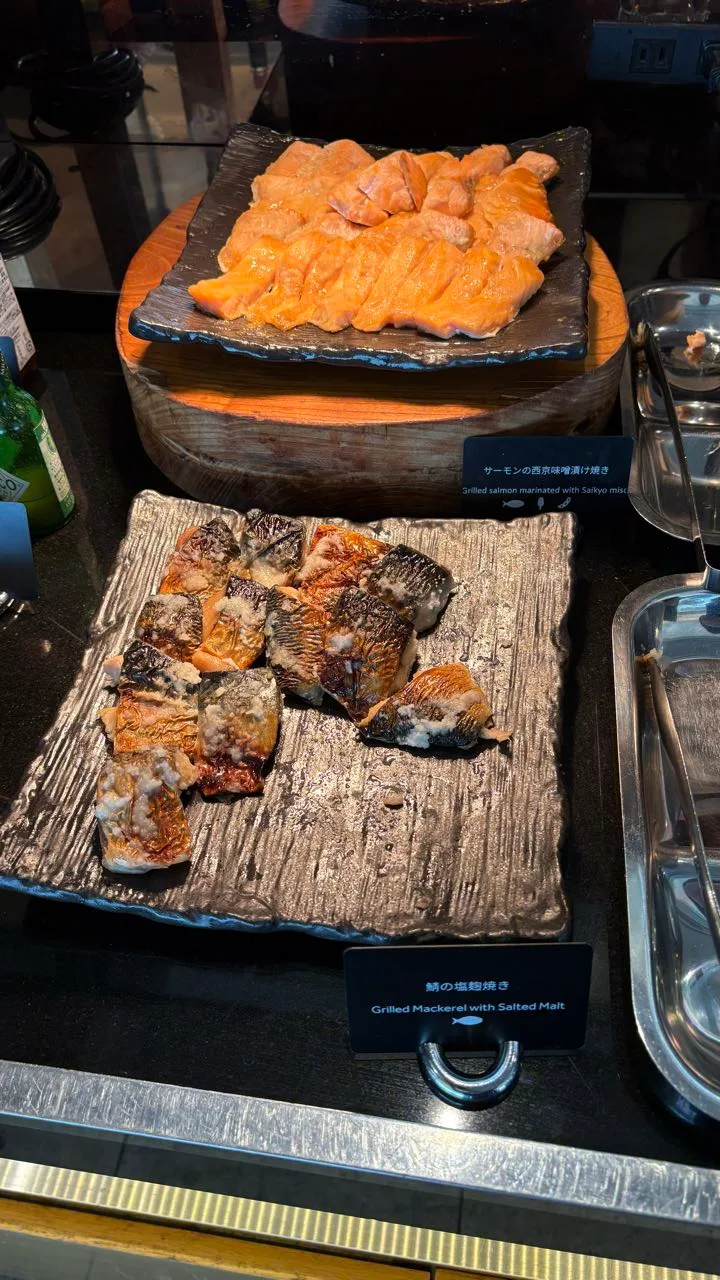 Two square ceramic plates in a warming counter, salmon fillets on the upper plate and pieces of grilled mackerel with charred skin on the lower one, each with a bilingual card