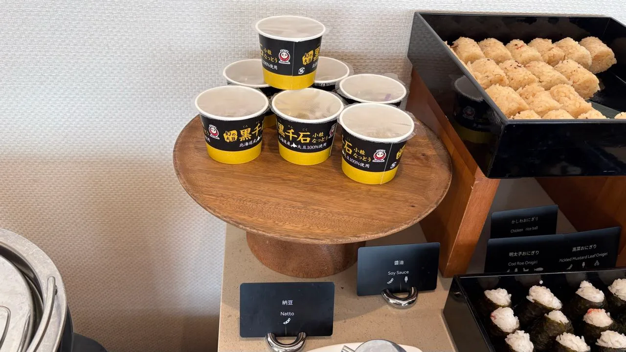 A wooden stand holding paper cups of natto with black and yellow labels, with trays of rice balls to the right and small black cards below reading Natto and Soy Sauce