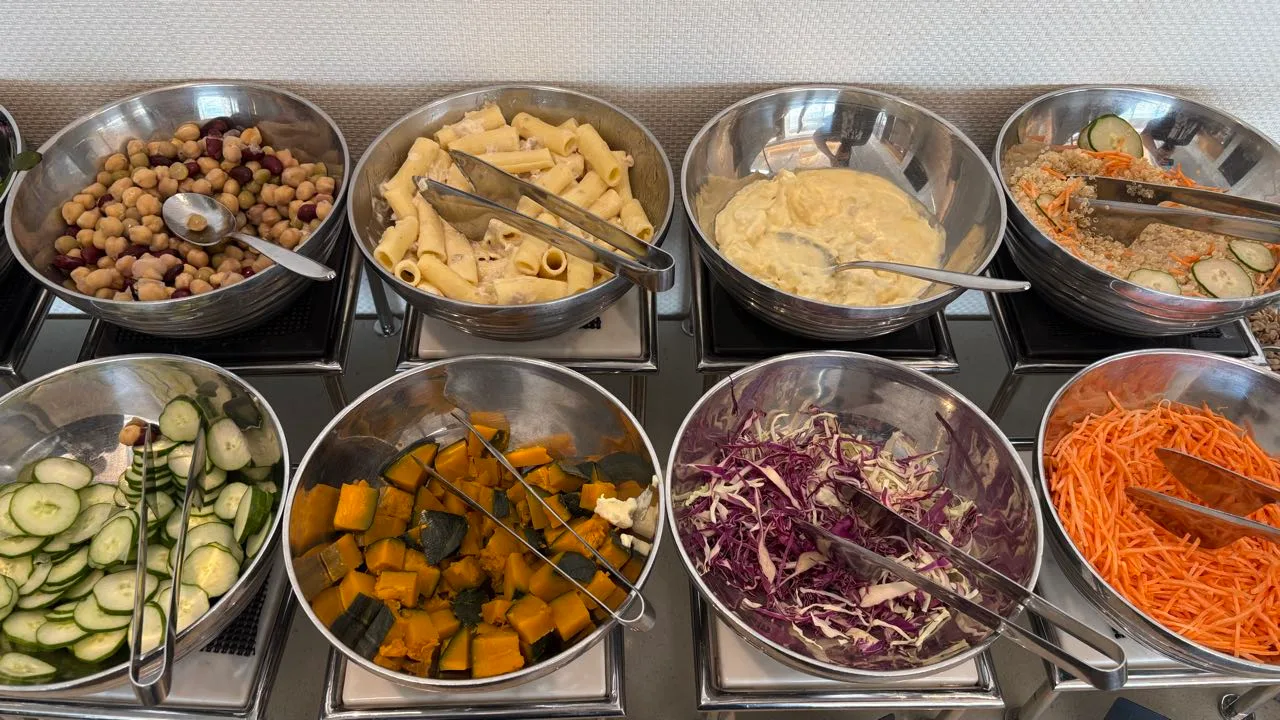 Eight stainless steel bowls in two rows holding mixed beans, pasta salad, potato salad, quinoa with cucumber and carrot, sliced cucumber, cubed pumpkin, shredded purple cabbage and grated carrot