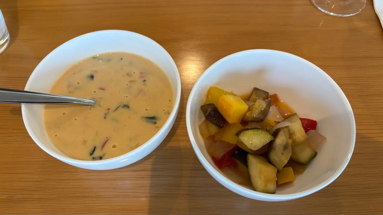 Two white bowls on a wooden table, one holding a creamy pale soup with a spoon in it, the other holding cubes of eggplant, zucchini, yellow and red peppers and onion
