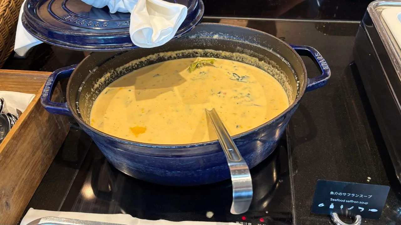 A blue cast-iron pot of thick pale yellow soup with a steel ladle in it, sitting on a black warmer, with a card beside it reading Seafood saffron soup