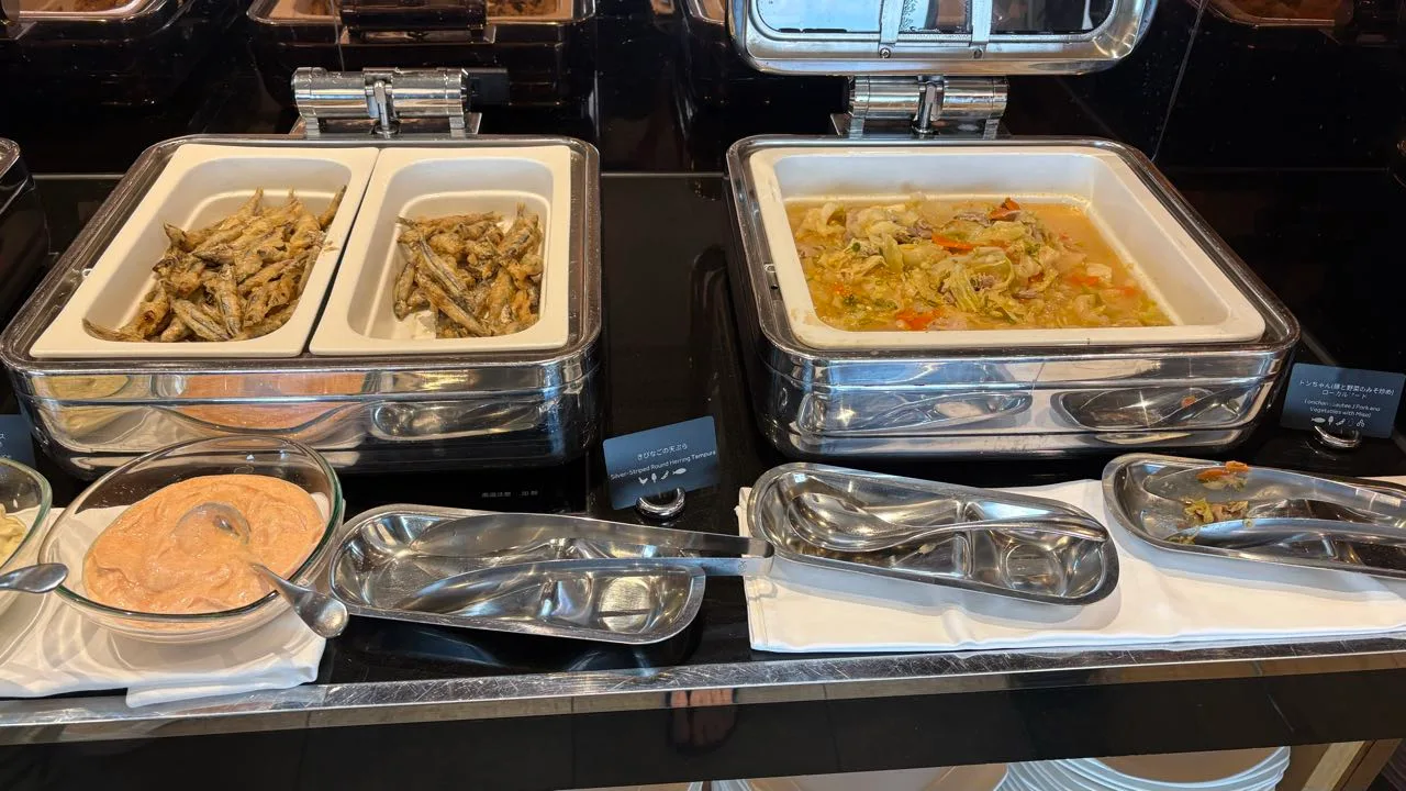 A buffet counter with two compartments of small fried fish on the left and a tray of pork and cabbage in broth on the right, with bilingual cards and a glass bowl of pink mayonnaise in front