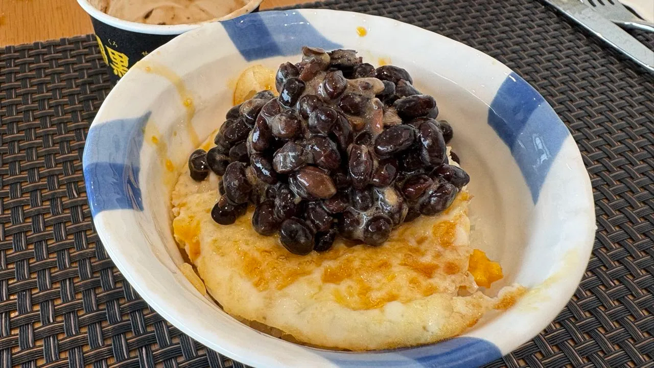 A blue and white bowl holding a fried egg over rice under a heaped pile of glossy dark natto beans, with the black natto cup behind it