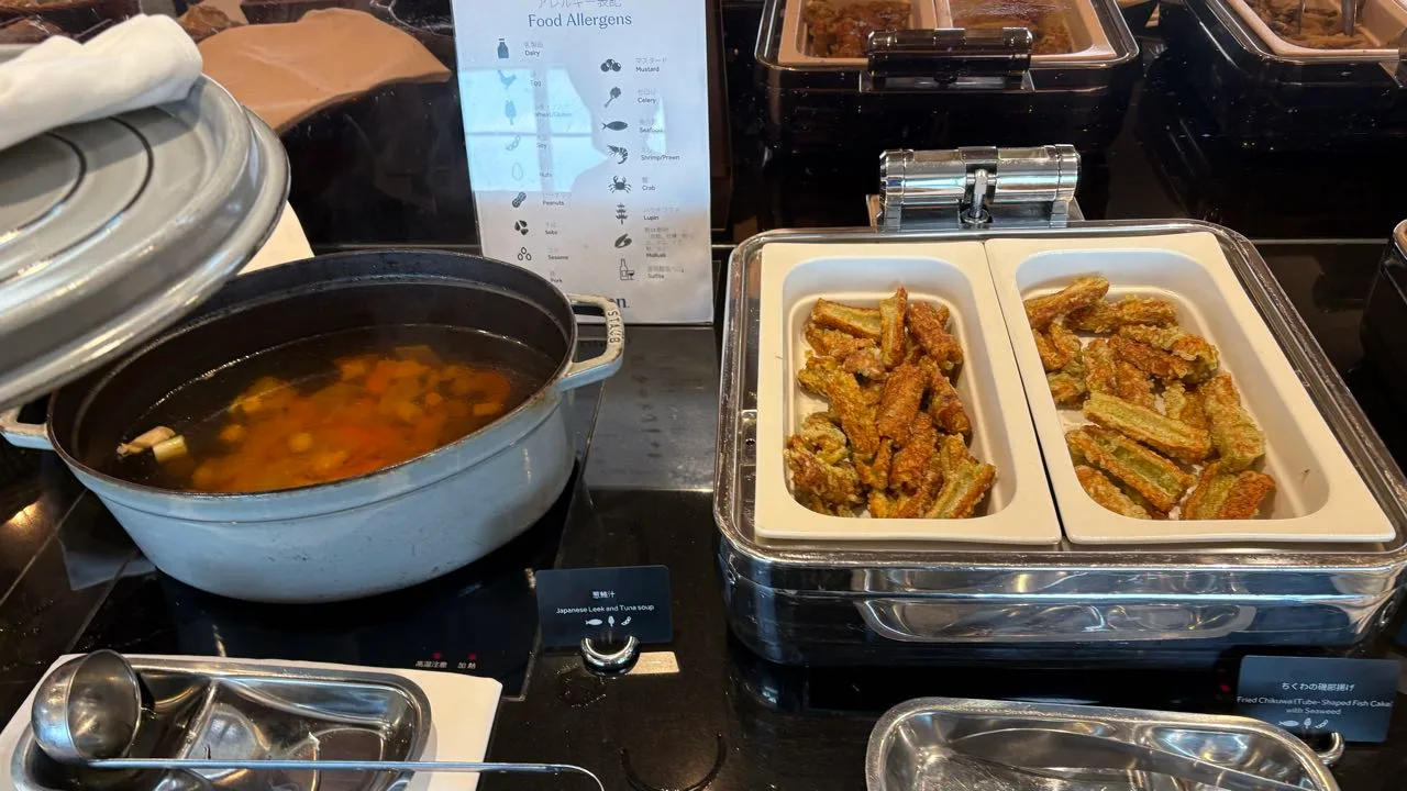 A pale blue enamel pot of clear soup with carrot and vegetables on an induction burner, a card reading Japanese Leek and Tuna soup, and a chafing dish of green-battered fried chikuwa beside it