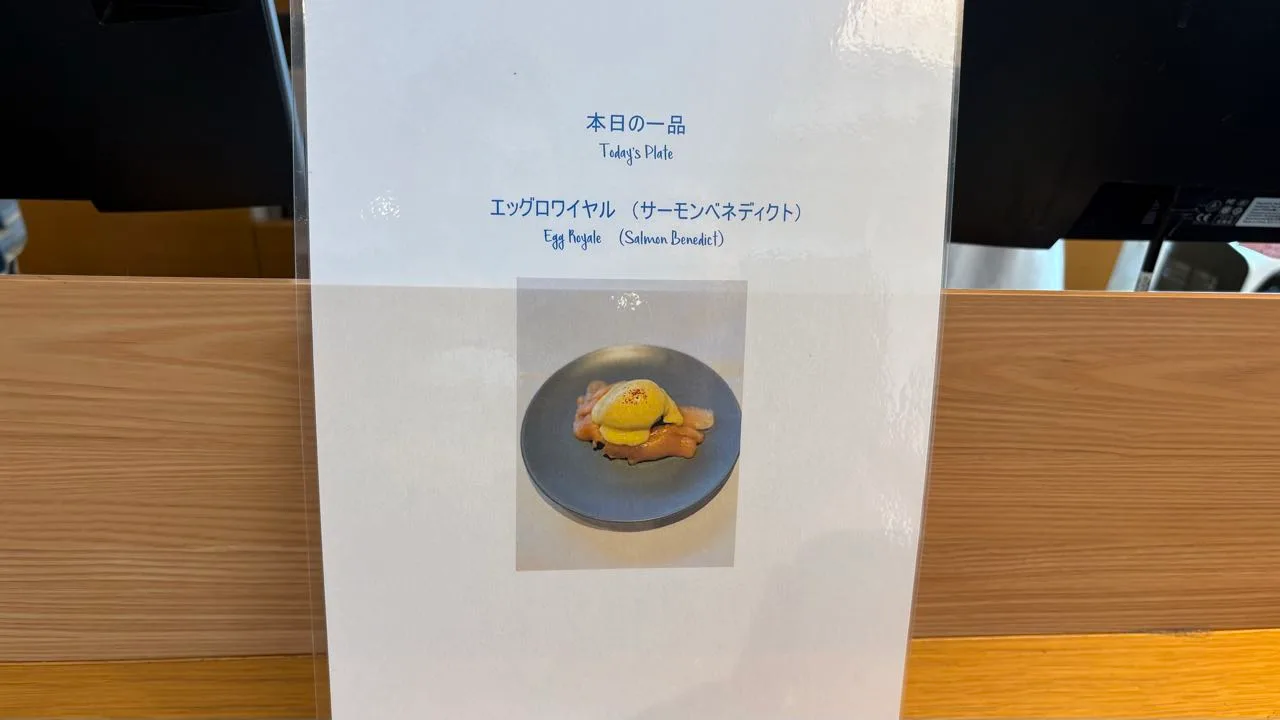 A laminated sign on a wooden counter reading Today's Plate and Egg Royale (Salmon Benedict) in Japanese and English, with a photograph of a poached egg under hollandaise on a grey plate