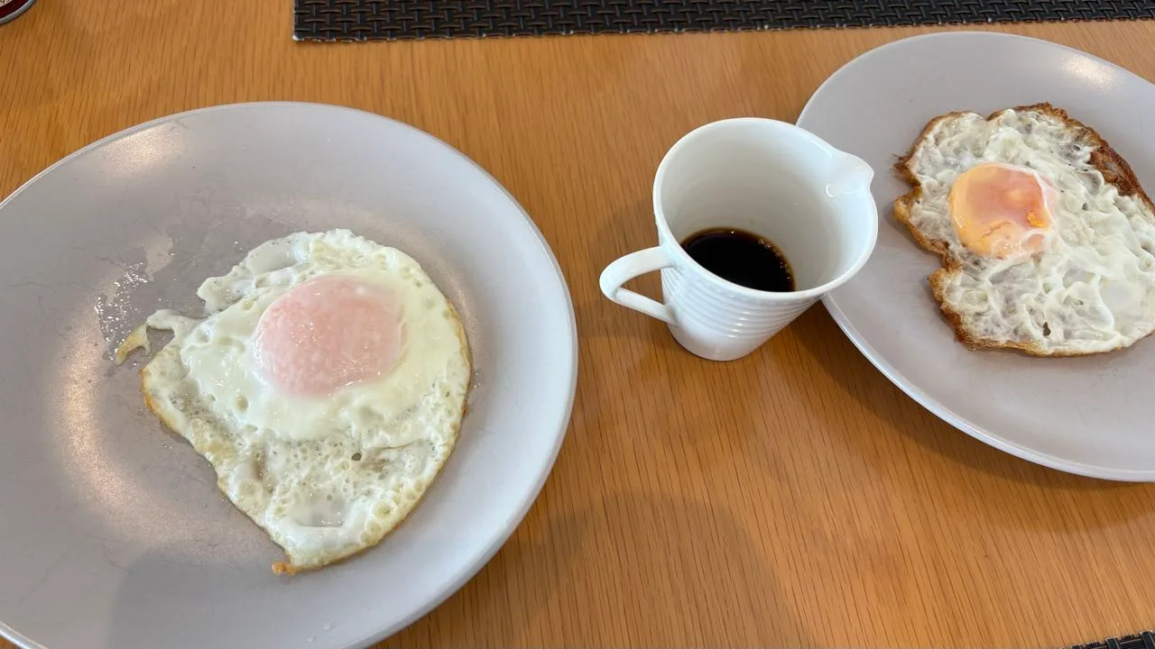 Two plates on a wooden table, the left holding a fried egg with a soft pink yolk and pale white, the right holding a fried egg with a browned crinkled white, a small white jug of dark sauce between them