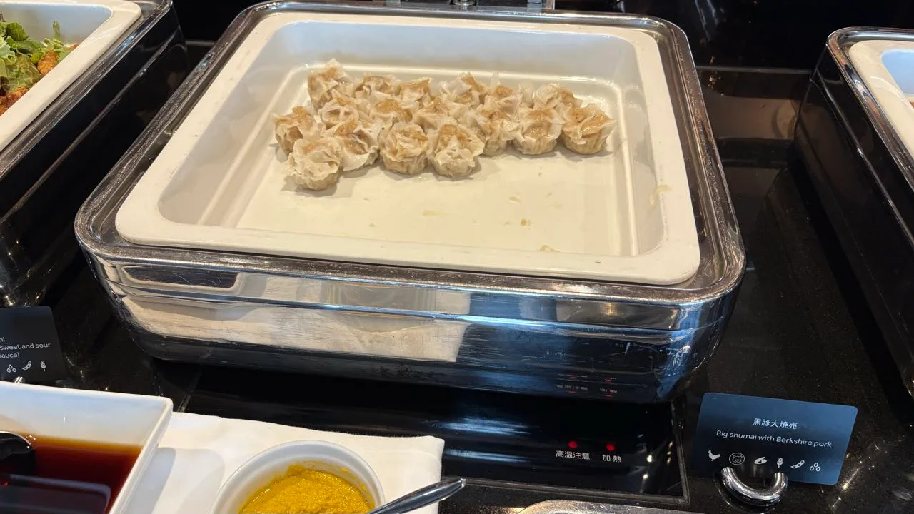 A chafing dish of shumai on a buffet counter with a card reading 黒豚大焼売 and Big shumai with Berkshire pork, with yellow mustard and soy sauce in dishes in front