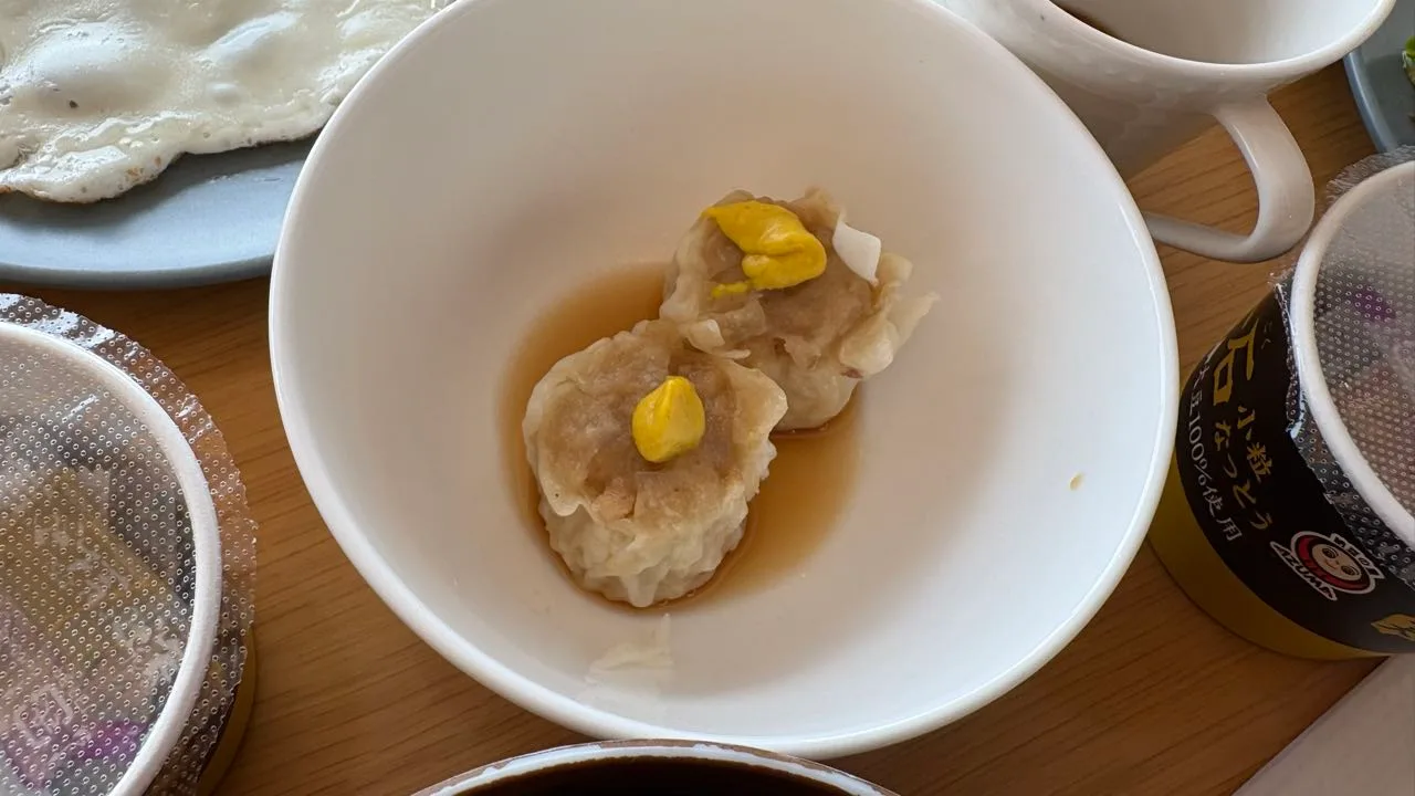 Two shumai sitting in broth in a white bowl, each topped with a dab of yellow mustard, with a fried egg on a grey plate behind and a natto cup at the right