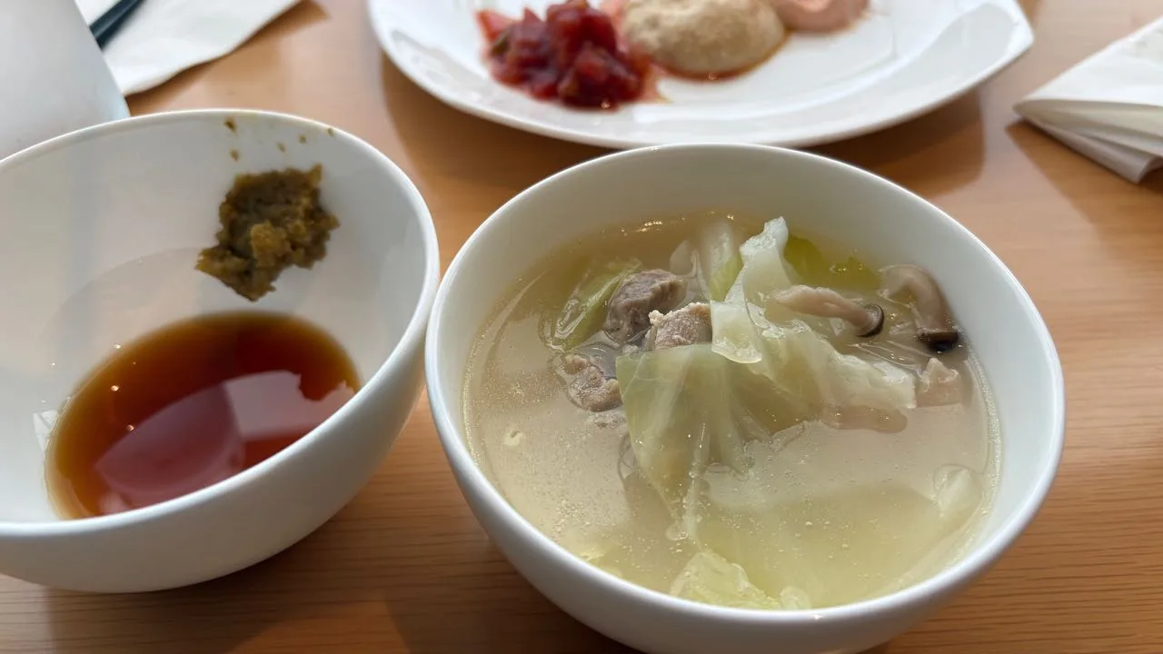 A white bowl of hot pot with cabbage, pieces of chicken and a mushroom in broth, beside a smaller white bowl holding brown ponzu and a dab of green yuzu kosho
