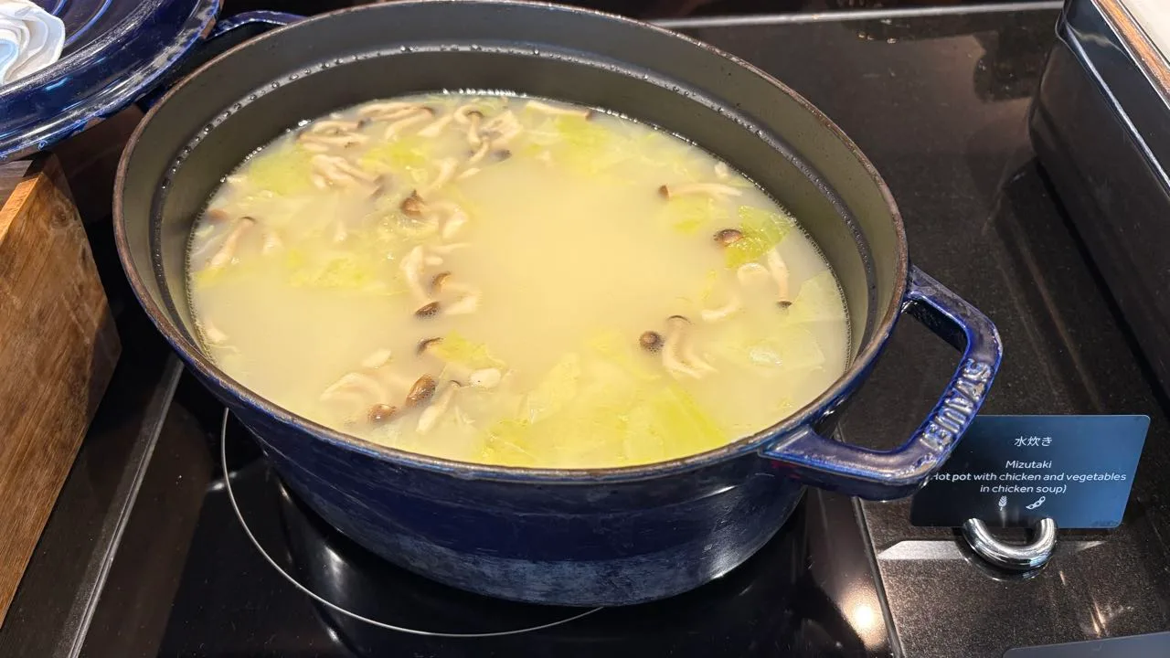 A blue cast-iron casserole on a hotplate holding pale broth with cabbage and shimeji mushrooms floating on the surface, and a card reading Mizutaki, hot pot with chicken and vegetables in chicken soup