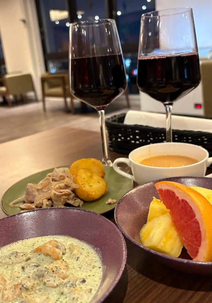 Two glasses of red wine with sliced pork and potatoes, a bowl of seafood in a green cream sauce, a cup of soup and a bowl of grapefruit and pineapple