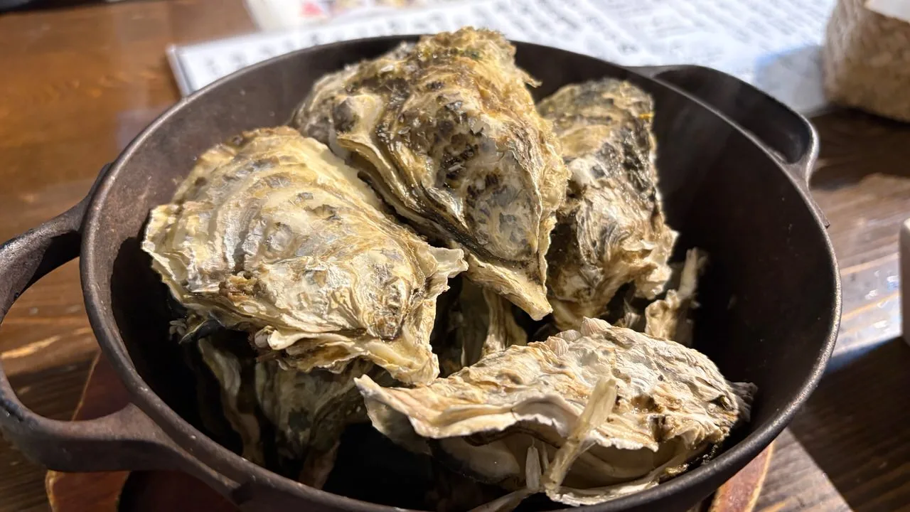 A black cast-iron pot filled to the rim with large steamed oysters in the shell on a wooden table