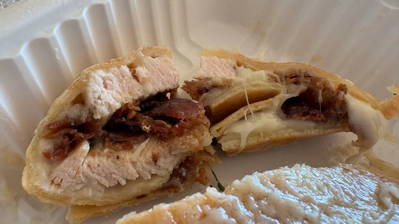 A chicken carbonara crepe cut in half in a takeout box, showing sliced chicken breast, dark crisp bacon and melted cheese inside