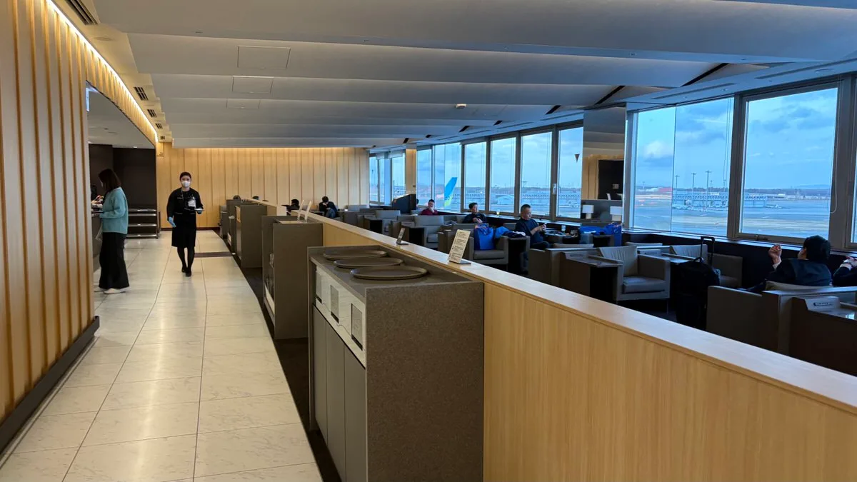 A long lounge room with pale wood panelling, armchairs facing a wall of windows over the airport apron, and a low counter down the middle