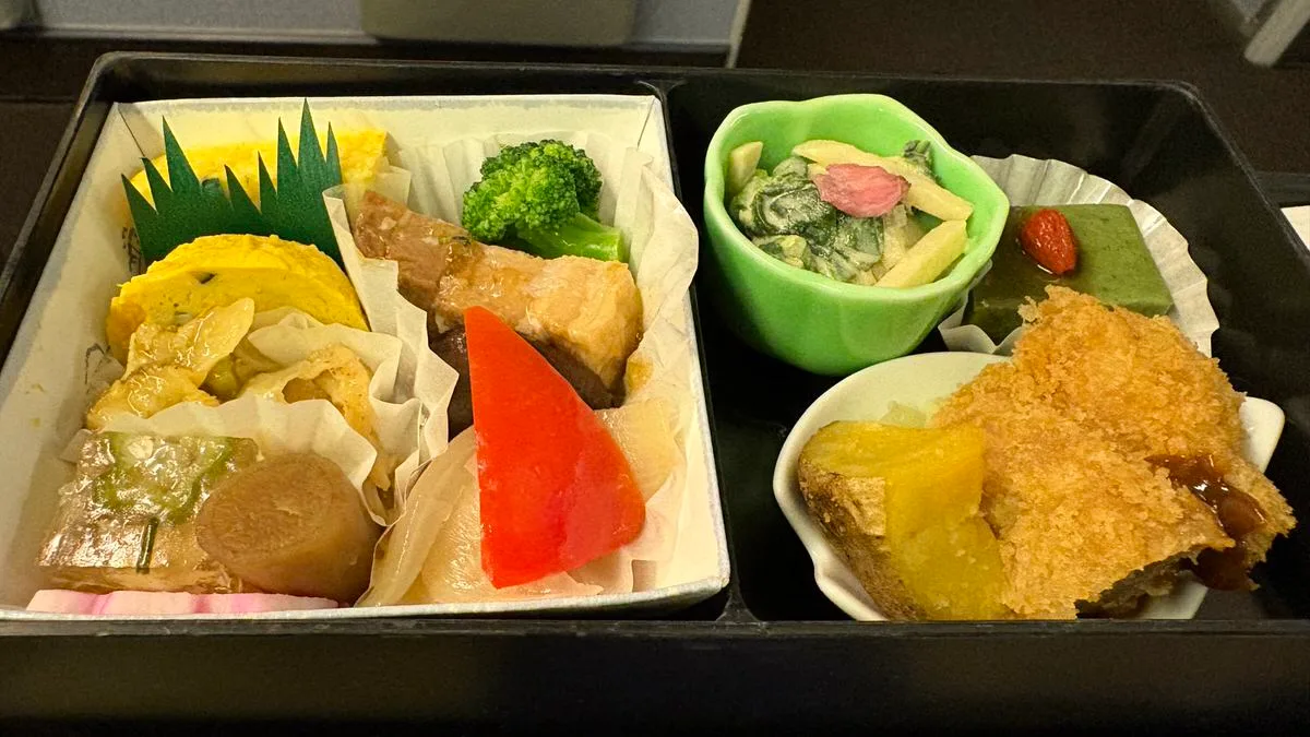 A black lacquer box divided into compartments holding rolled omelette, simmered pork, broccoli, a red carrot cut, a green bowl of dressed greens, a green cube of tofu and a fried cutlet with sauce