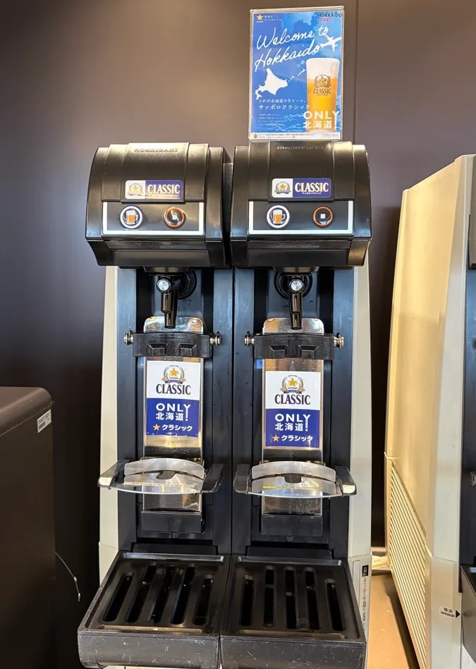 Two black automatic beer-pouring machines side by side, labelled Sapporo Classic, with a Welcome to Hokkaido poster above them