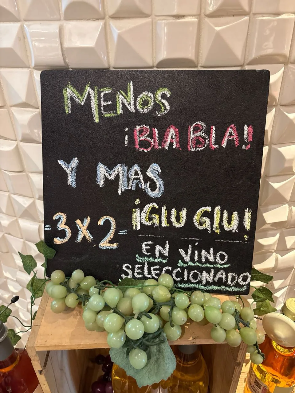 A chalkboard reading menos bla bla y mas glu glu, 3x2 en vino seleccionado, propped above bottles and plastic grapes