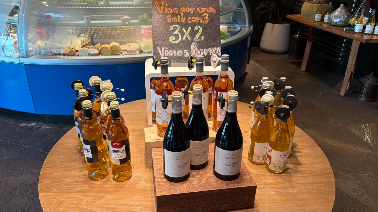A round wooden table set with white and rosé wine bottles under a chalkboard reading 3x2