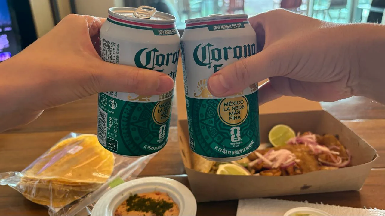 Two hands touching cans of Corona Extra together over a table, with a bag of tostadas, a bowl of dip and an open box of tacos behind
