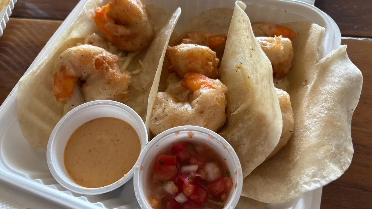 Three thin flour tortillas with battered shrimp in a white takeout box, next to a cup of orange creamy sauce and a cup of pico de gallo