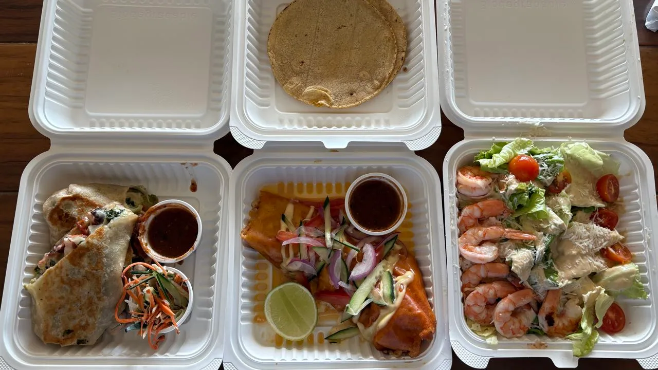 Three open takeout boxes: a burrito with salsa and a cup of carrot and cucumber slaw, a shrimp costra with lime and corn tortillas in the lid, and a shrimp Caesar salad