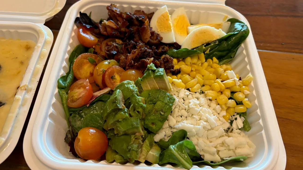 A takeout box of spinach topped with bacon bits, boiled egg wedges, corn, avocado, cherry tomatoes and crumbled white cheese