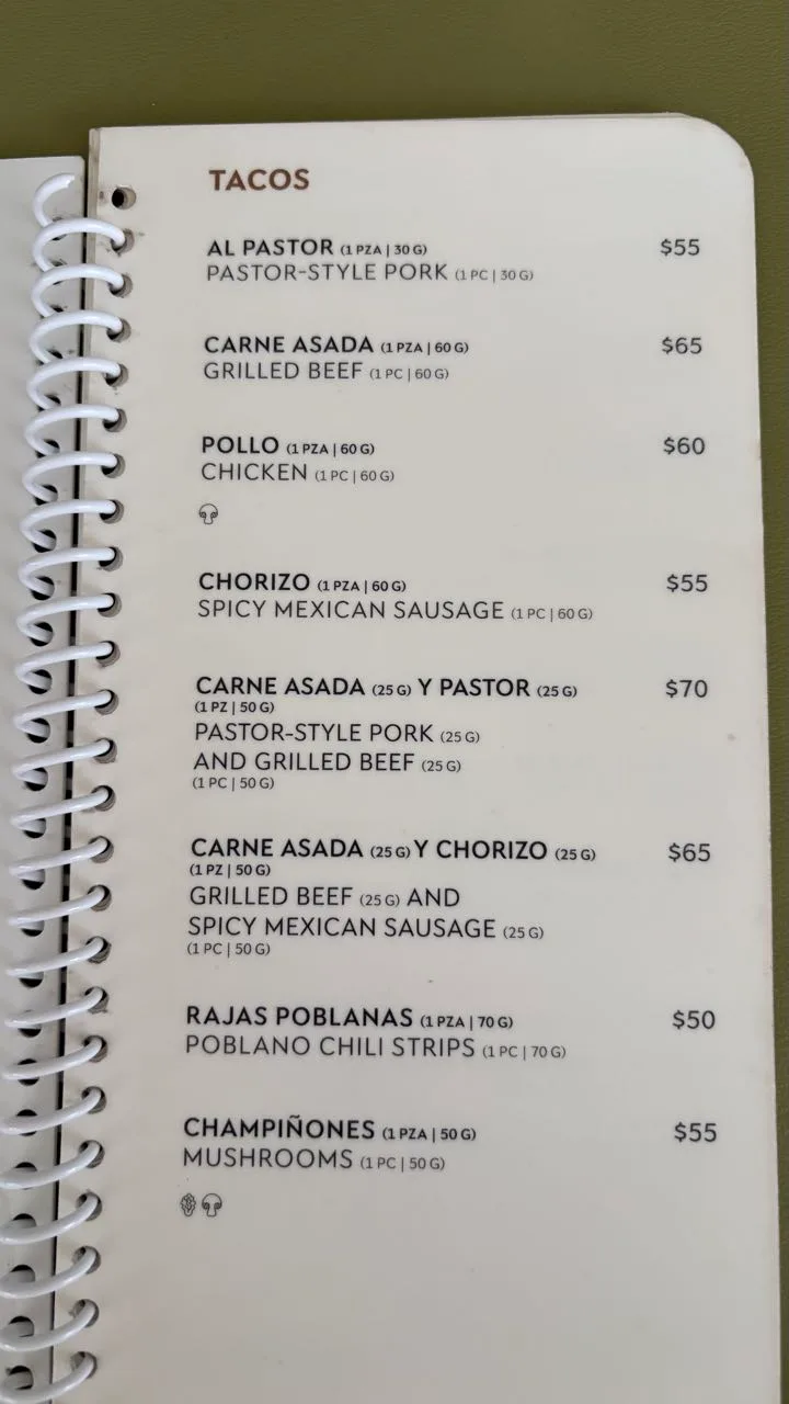 A spiral-bound menu page headed TACOS listing eight single tacos in Spanish with English below, priced 50 to 70